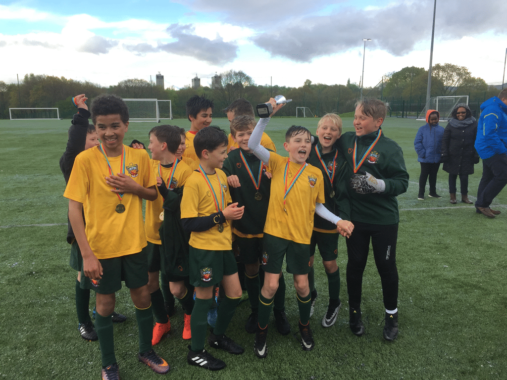 cheadle hulme school year 7 U12 football team raise their cup winners trophy on the pitch wearing their football kit as they celebrate after winning the stockport schools football league